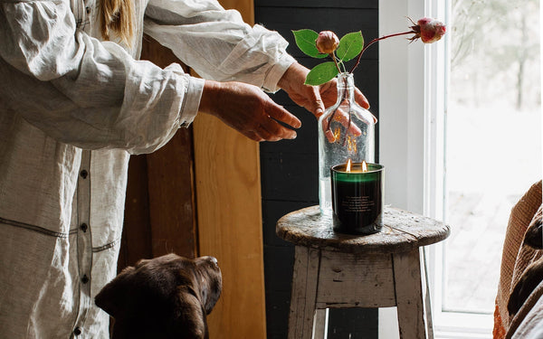 Southern Wild Co scented candle burning beside a rose stem in a glass bottle, styled on a rustic wooden stool. A woman in a linen smock arranges flowers while a chocolate Labrador watches. Natural light, slow living, and Australian-made home fragrance.