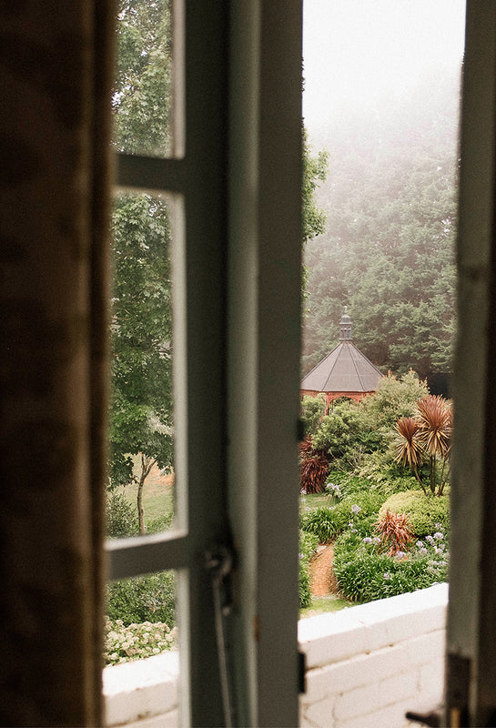 View through a cottage window onto a misty autumn garden with trees and an old gazebo, capturing the quiet atmosphere of a cool country morning.