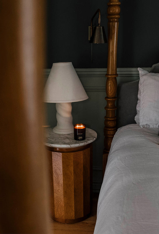 Southern Wild Co Our Place candle burning on a bedside table beside linen bedding and soft lamplight, creating a warm and restful autumn bedroom scene.