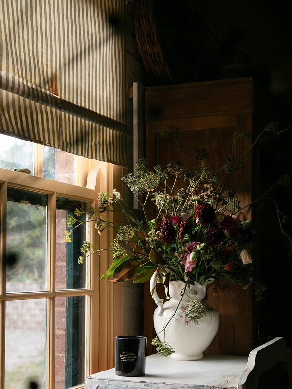 Bush Magic candle nestled beside a wild bouquet in a white ceramic jug, catching the soft window light of a rustic country home.
