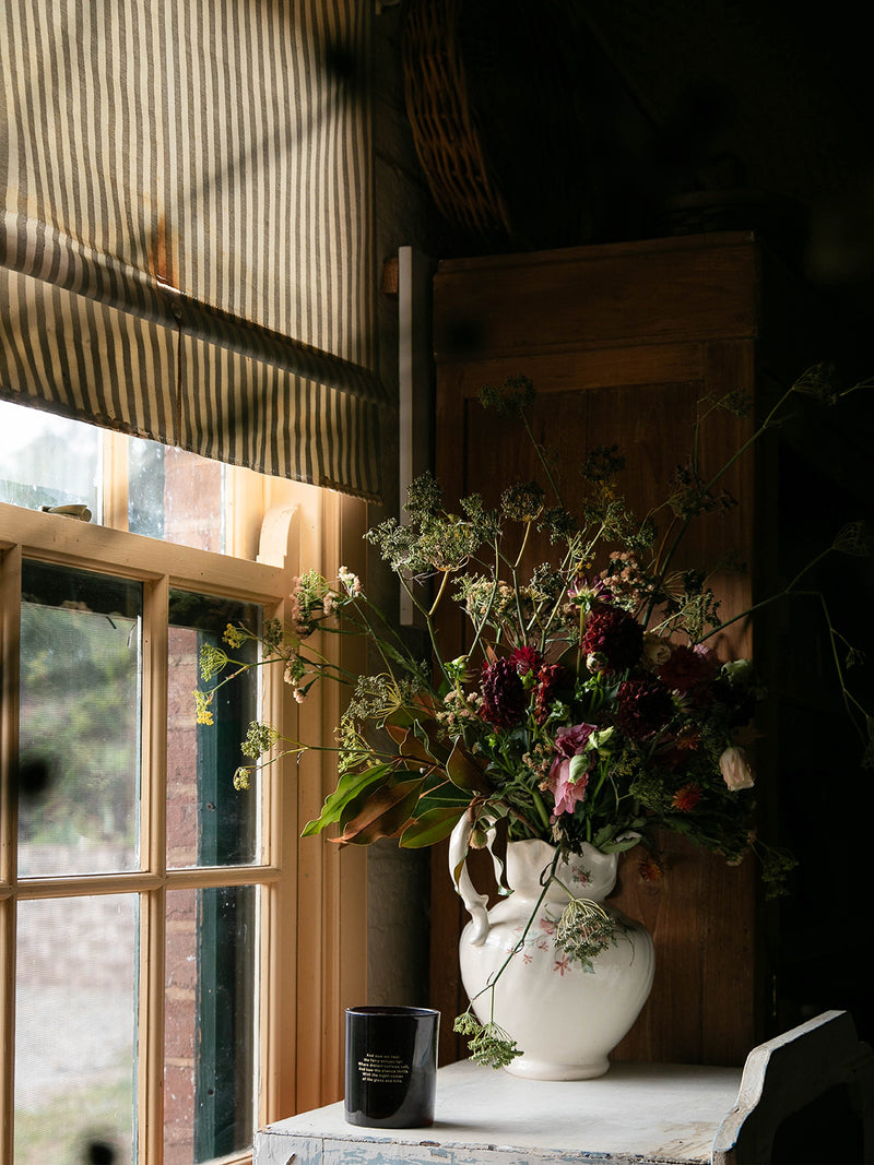 Bush Magic candle nestled beside a wild bouquet in a white ceramic jug, catching the soft window light of a rustic country home.
