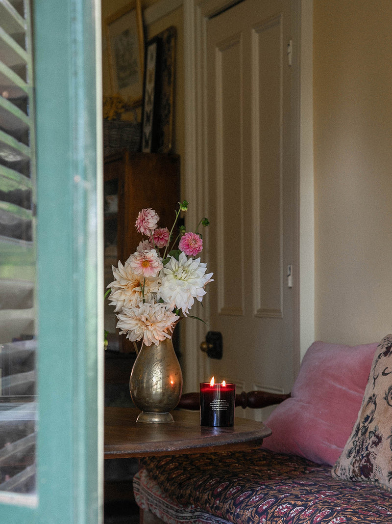 Bush Magic candle casting a warm glow on a vintage table in a cosy heritage room, paired with dahlias in an antique brass vase and soft velvet cushions.