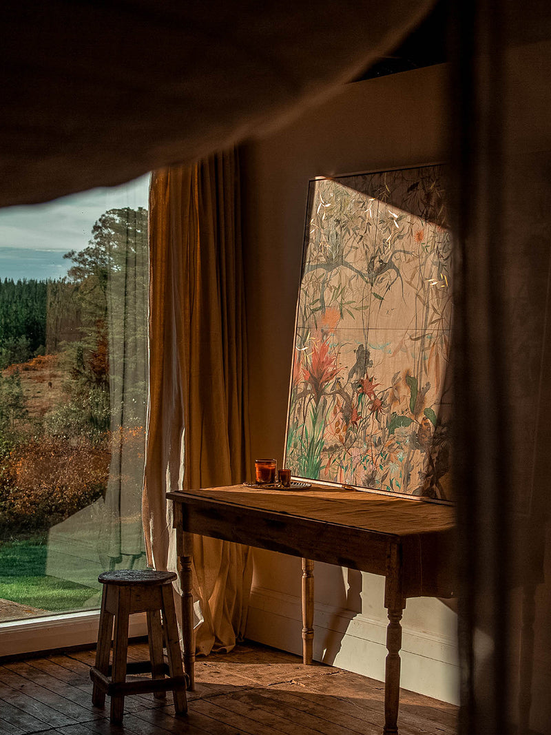 Bush Magic candle resting on a timber table near botanical artwork and soft golden light, with sweeping views to the wild Australian bush beyond.