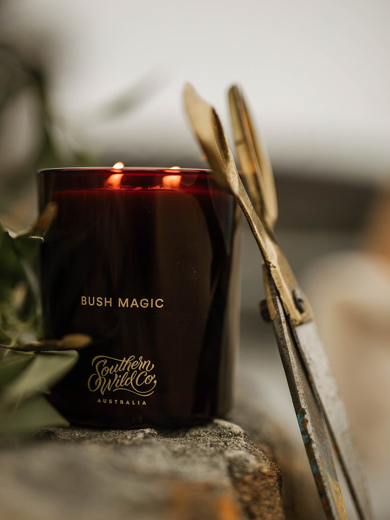 Bush Magic candle glowing with twin flames beside antique brass scissors on a stone ledge, evoking the rugged romance of wildcraft and ritual.