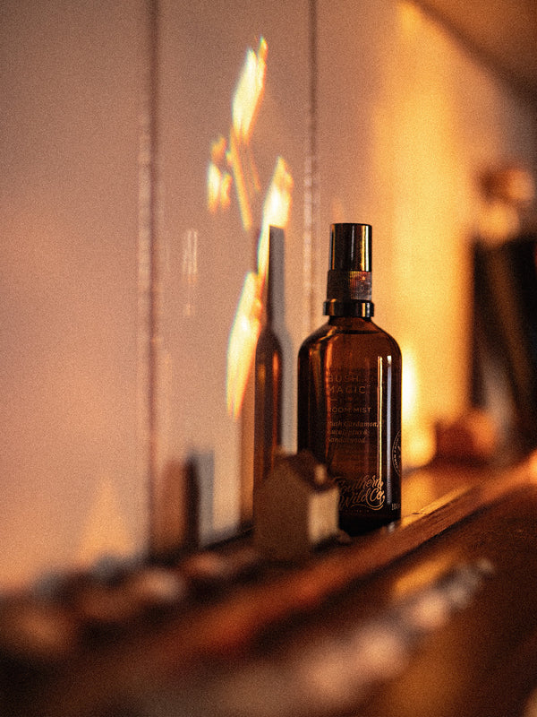 Bush Magic Room Mist bathed in the golden glow of sunset, resting on a wooden ledge with reflected light dancing on the wall like fire and foliage.