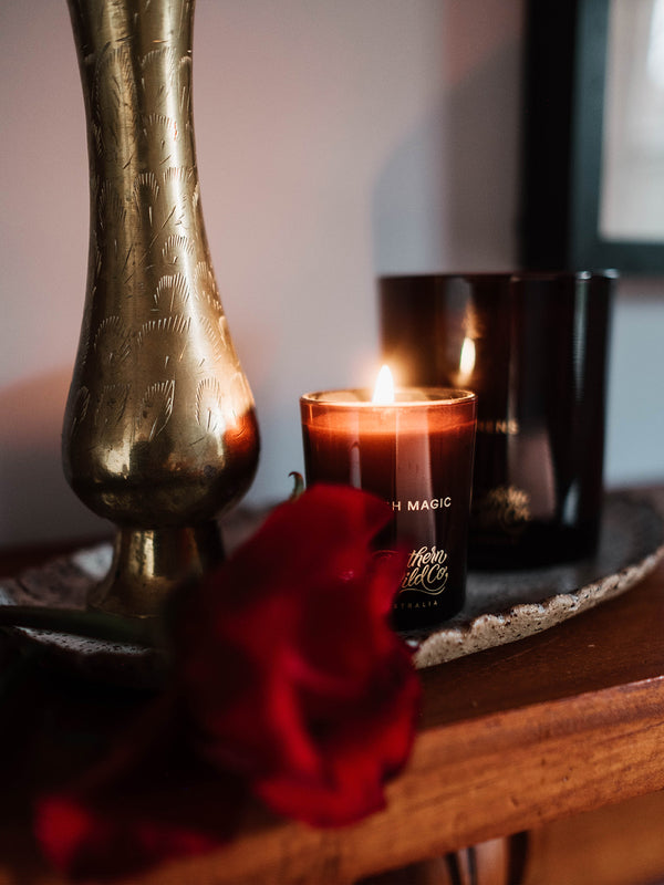 Bush Magic votive candle flickering beside a vintage brass vase and red rose, conjuring an atmosphere of bushland romance and moody stillness.