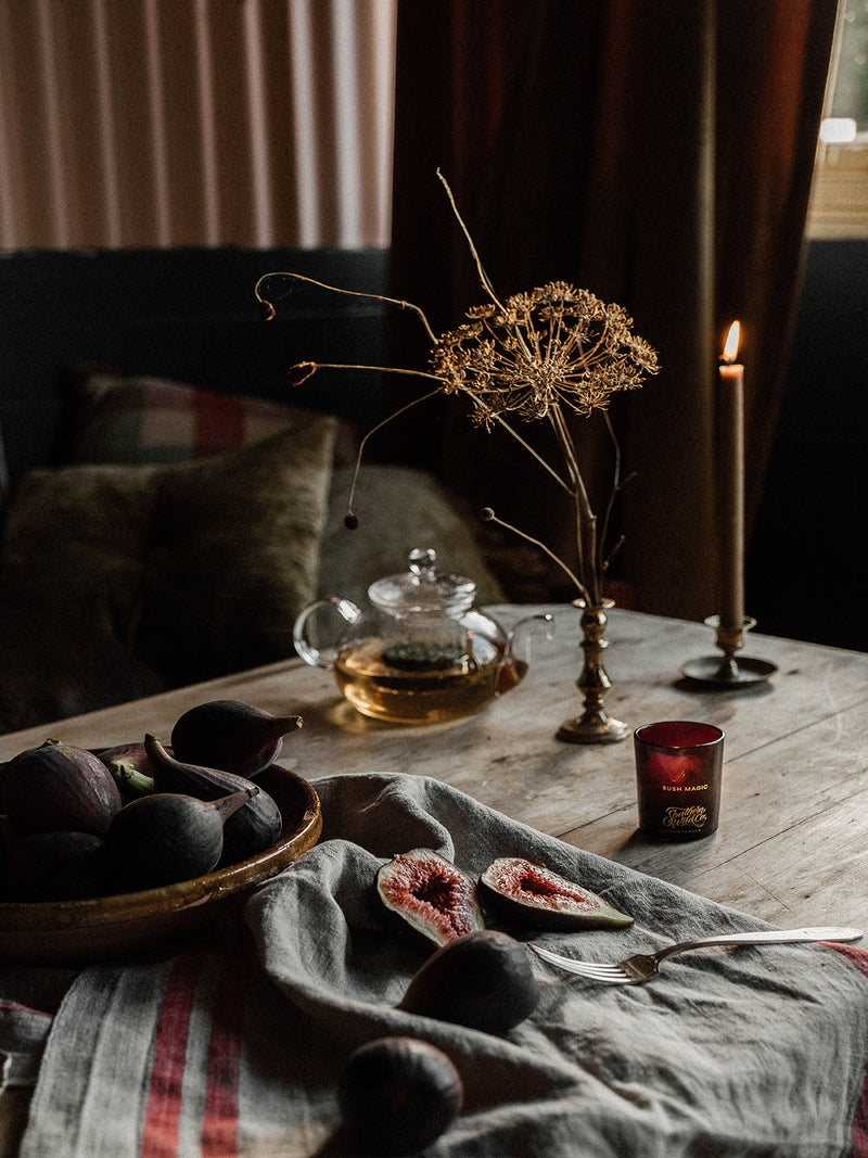 A quiet still life of figs, herbal tea, and the glowing Bush Magic votive candle on linen-draped timber – a scene of rustic sensuality and calm.