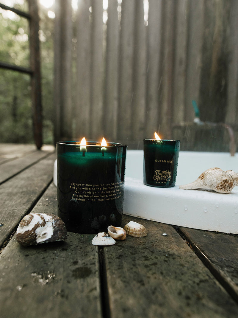 Ocean Isle candles—votive and large—set beside seashells on a rain-dampened deck, capturing the scent and serenity of an outdoor soak by the sea.