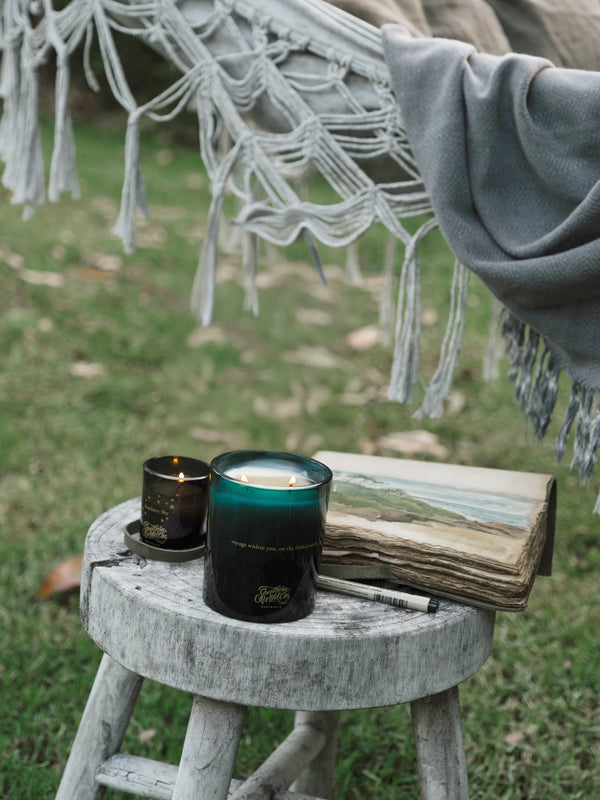 Ocean Isle candles beside a weathered sketchbook and hammock fringe, inviting moments of reflection in a quiet garden beneath a soft afternoon sky.
