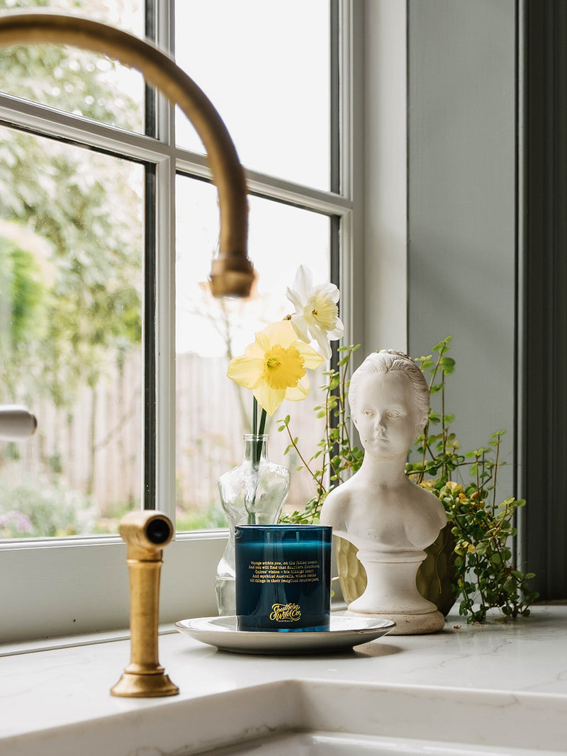 Ocean Isle candle placed on a dish beside daffodils in a glass bottle and a classical bust statue on a kitchen windowsill—sunlight and scent mingling in a quiet morning moment.