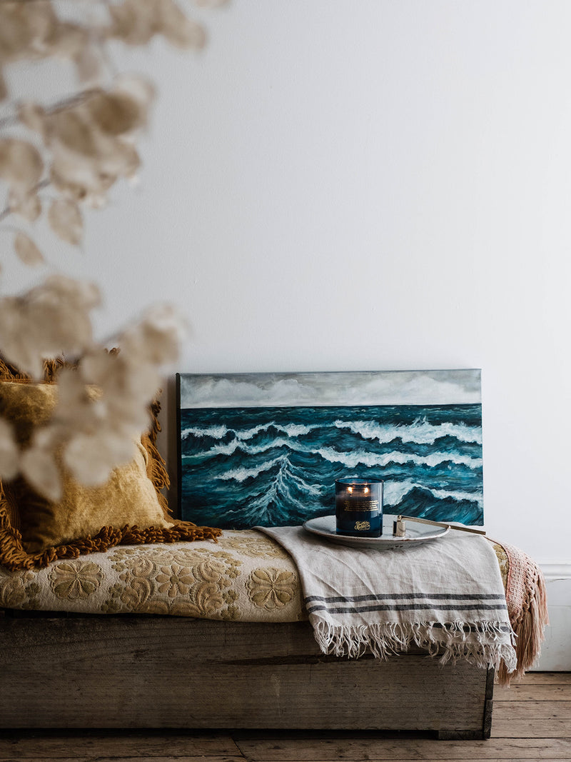 Ocean Isle candle resting before a seascape painting on a vintage daybed layered with soft linen and golden cushions—bringing scent and stillness to a coastal-inspired nook.