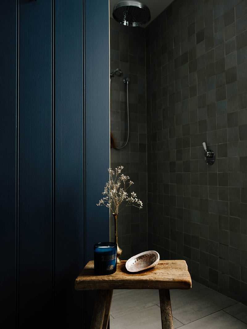 Ocean Isle candle beside a weathered timber stool, abalone shell and dried flowers creating a quiet ritual space in a moody tiled bathroom.