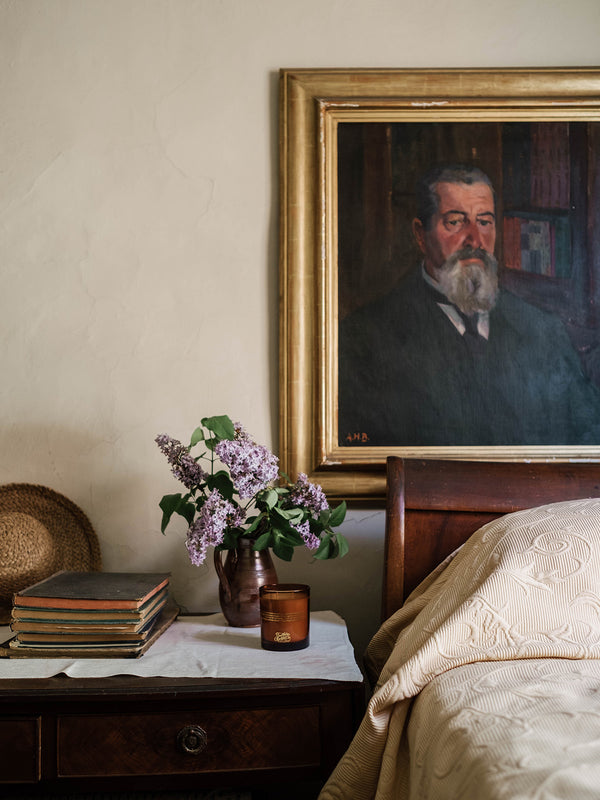 Our Place candle by Southern Wild Co styled on a timber bedside table with vintage books, lilacs, and an antique portrait – Australian made candle for heritage bedrooms and layered interiors.