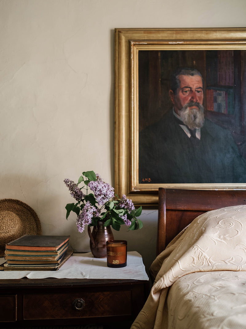 Our Place candle by Southern Wild Co styled on a timber bedside table with vintage books, lilacs, and an antique portrait – Australian made candle for heritage bedrooms and layered interiors.