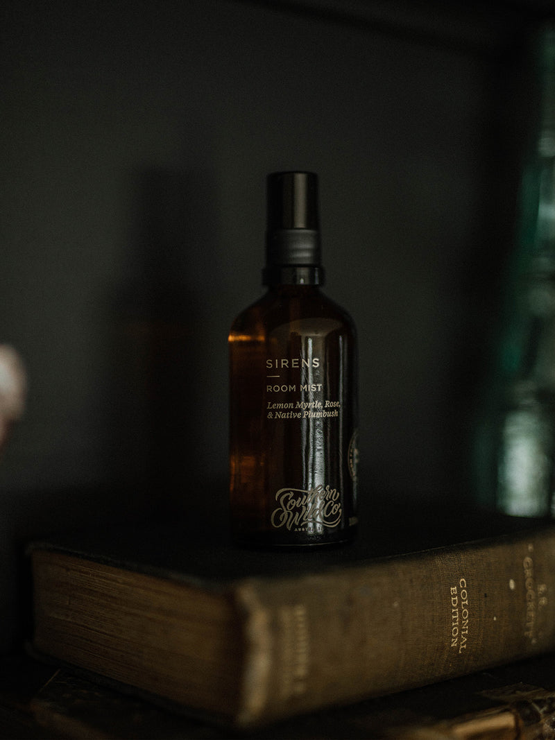 Amber glass Sirens Room Mist bottle resting on worn book spines, with soft morning light evoking history, scent and storytelling.