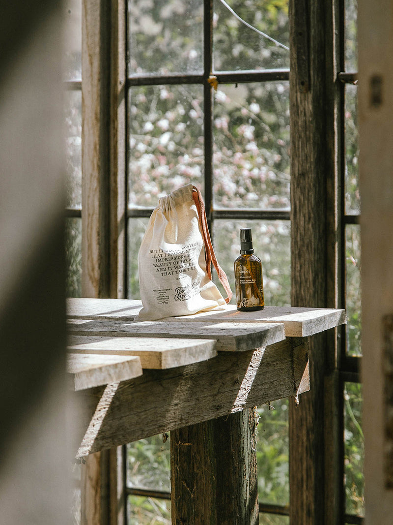 Sirens Room Mist with linen drawstring pouch resting on a weathered timber table, sunlight filtering through vintage windows and soft spring blooms beyond.