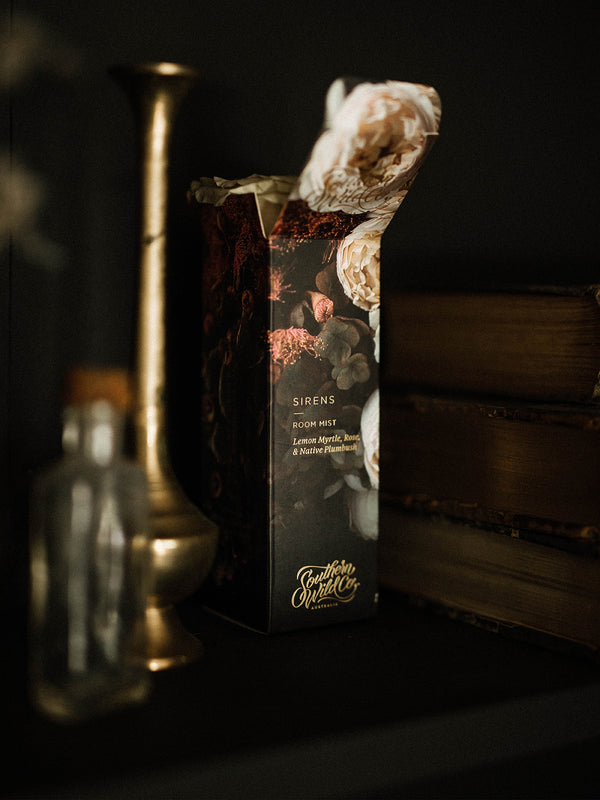 Beautifully boxed Sirens Room Mist tucked among antique brass, old books and shadows — a quiet moment of scent and stillness.
