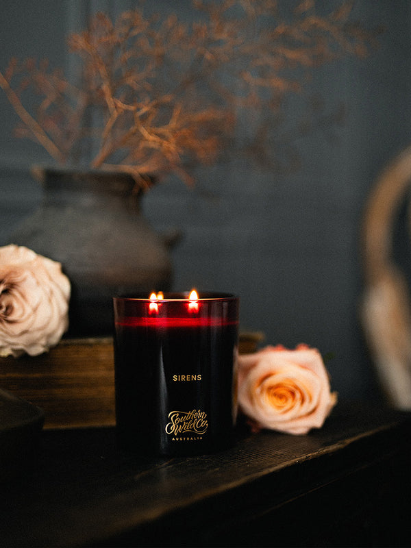 Sirens candle by Southern Wild Co burning softly beside blush roses and earthy branches, evoking a quiet, poetic stillness in a moody interior.