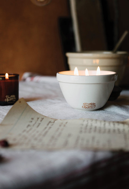 Two lit Southern Wild Co Magic Pudding candles ion a table with a vintage recipe and a blurred background