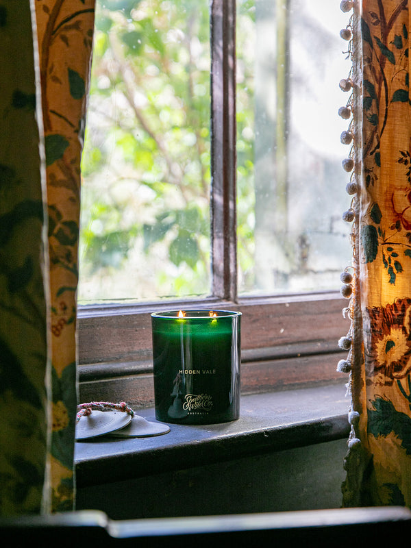 Southern Wild Co Hidden Vale candle glowing on an old timber windowsill framed by vintage floral curtains – Australian made candle for heritage homes and bush-inspired interiors.