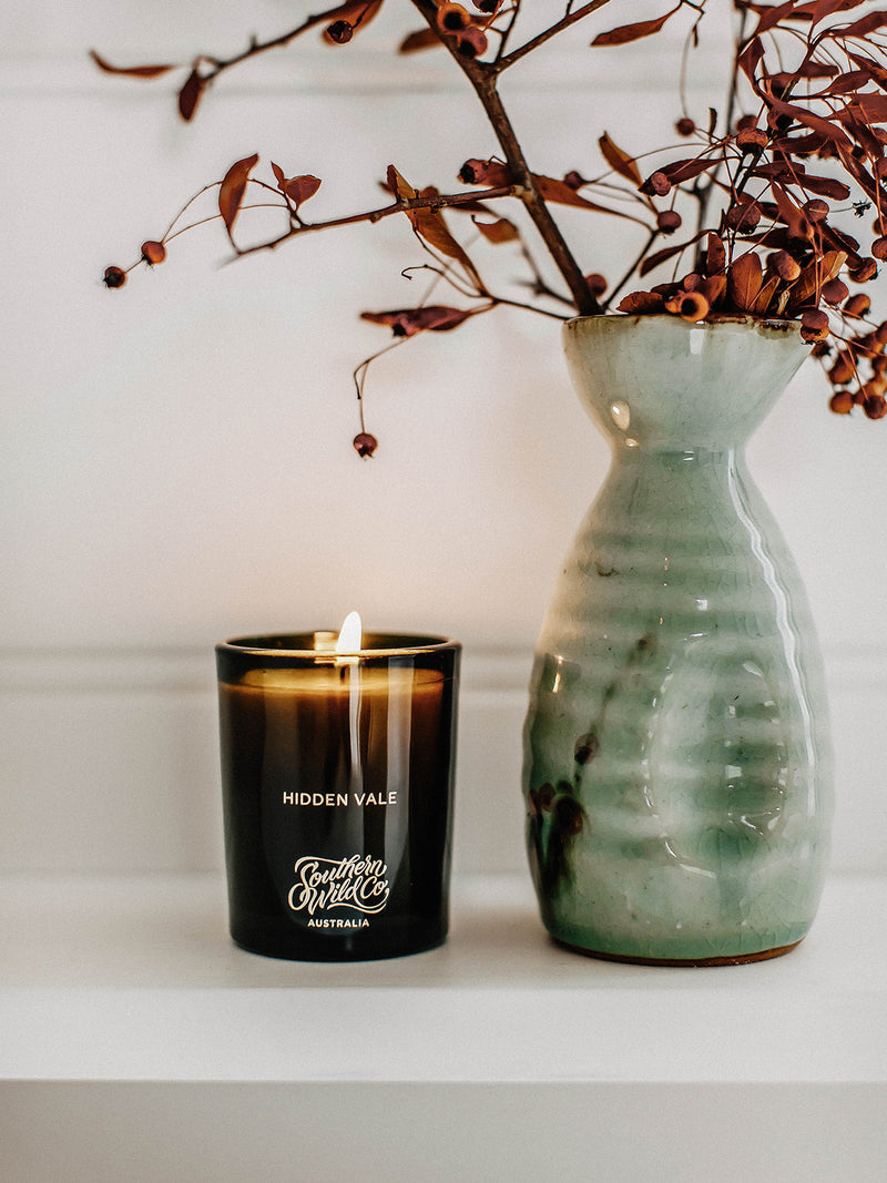 Hidden Vale soy candle by Southern Wild Co burning beside a green ceramic vase with dried native Australian botanicals on a white shelf – Australian made candle for calming rituals and interior styling.