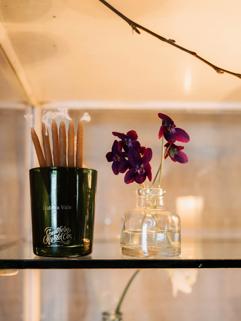 Repurposed Hidden Vale candle vessel by Southern Wild Co used to hold beeswax taper candles, styled beside a glass jar of violets – sustainable candle packaging with a touch of country charm.