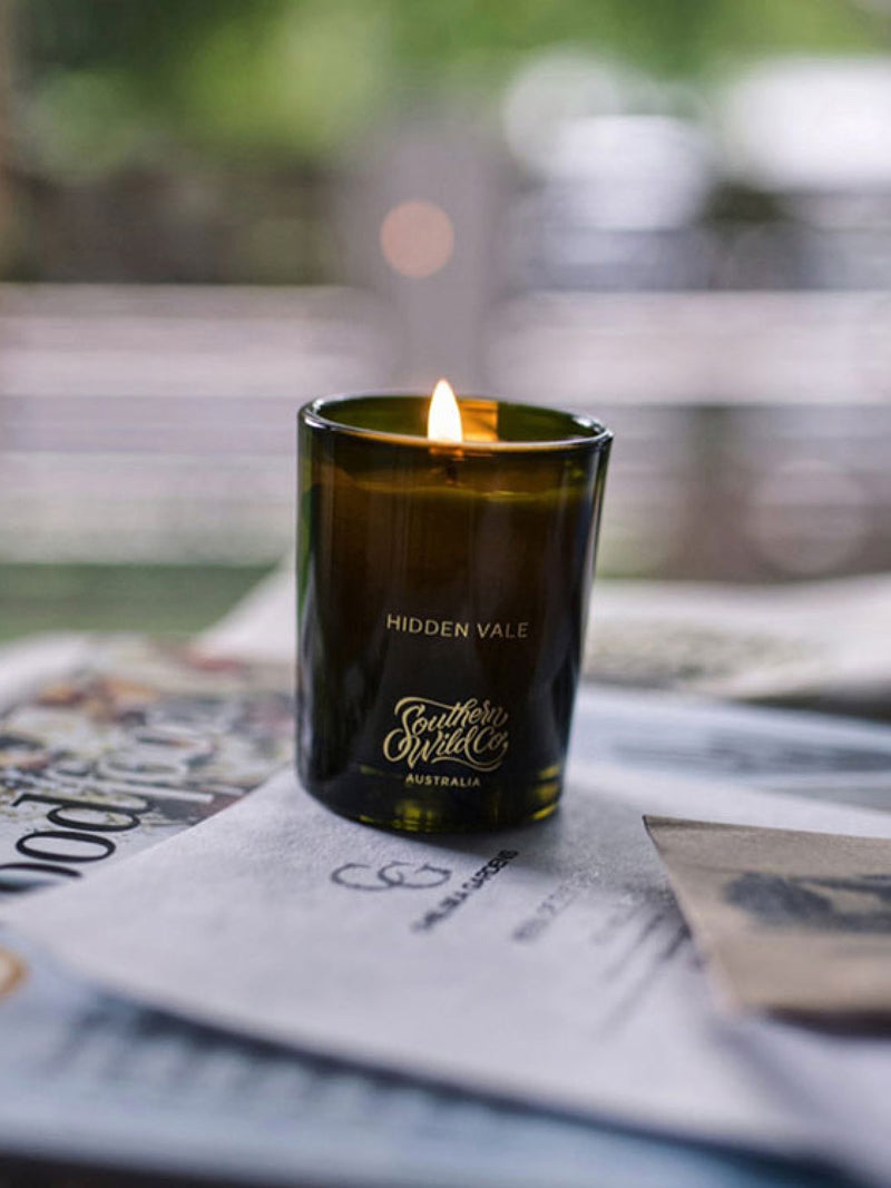 Hidden Vale candle by Southern Wild Co glowing on a table of books and paper in a peaceful indoor setting – premium Australian soy wax candle for home sanctuary and creative spaces.