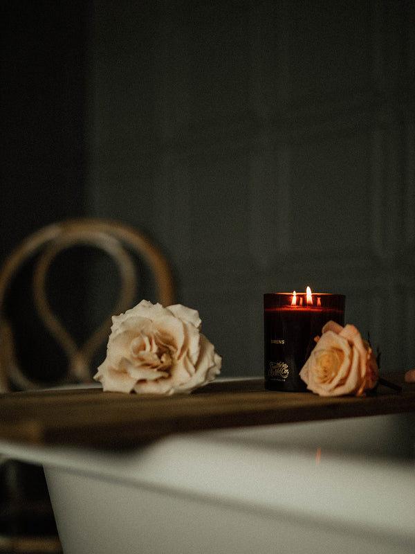 Sirens votive candle by Southern Wild Co glowing beside pale garden roses on a wooden bath tray – a quiet ritual of scent, softness, and self-care.
