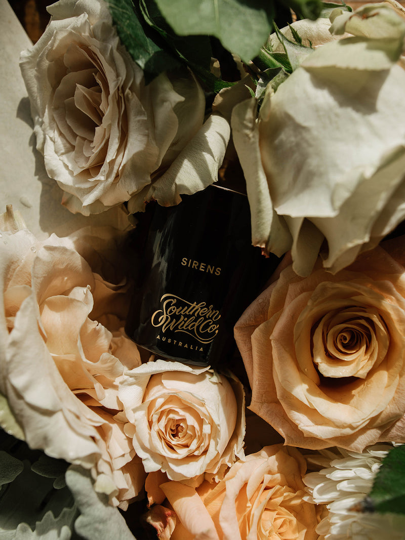 Southern Wild Co’s Sirens candle glowing against dark moody tones, softened with dried branches and blush roses – evocative and romantic.