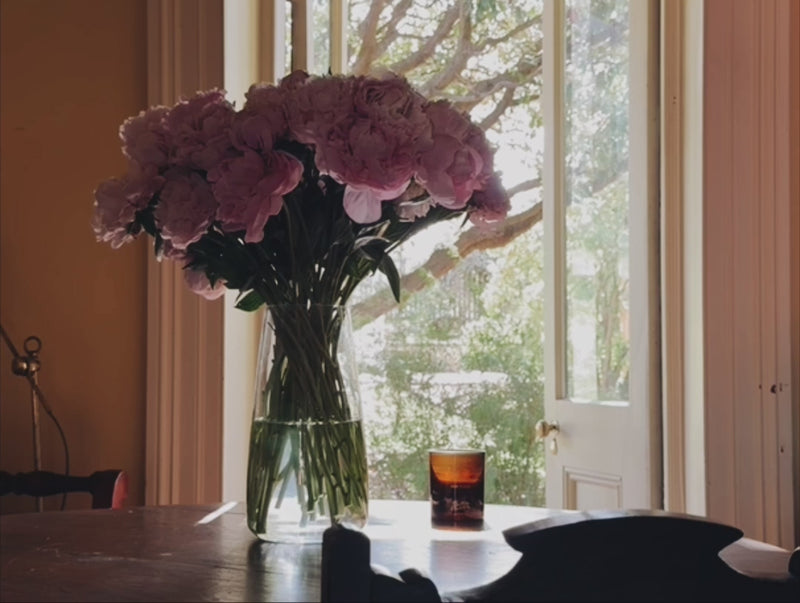 A tranquil scene of peonies in a vase looking through to a sunlit garden. There is a Southern Wild Co candle on the table
