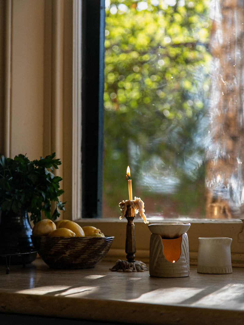 Clay Beehive Oil Burner glowing on a sunlit windowsill beside a brass candle holder and bowl of lemons – a quiet home ritual by Southern Wild Co.