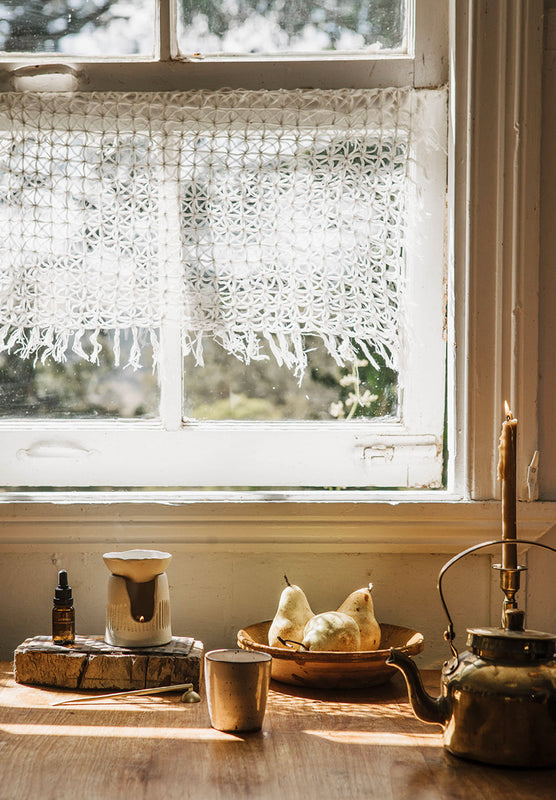 A rustic white crochet lace curtain with fringe hangs over a vintage window in a sunlit room. A wooden table below holds a metal mug, a small ceramic vessel, a bowl of pears, and a brass kettle.