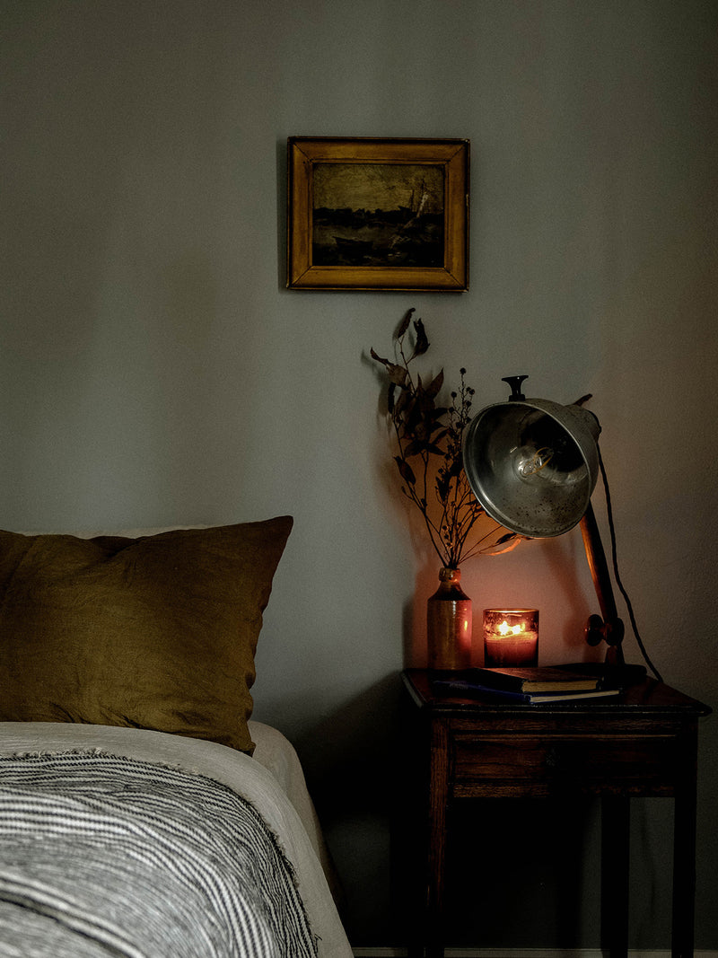 A Southern Sky candle glows softly on a vintage bedside table beneath a warm-toned painting and dried native branches, casting a golden light over a peaceful, earthy bedroom.