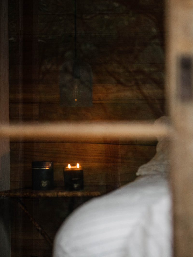 Seen through a window, the soft twin flames of the Southern Sky candle burn gently inside a timber-lined cabin—inviting warmth, stillness, and deep night dreaming.