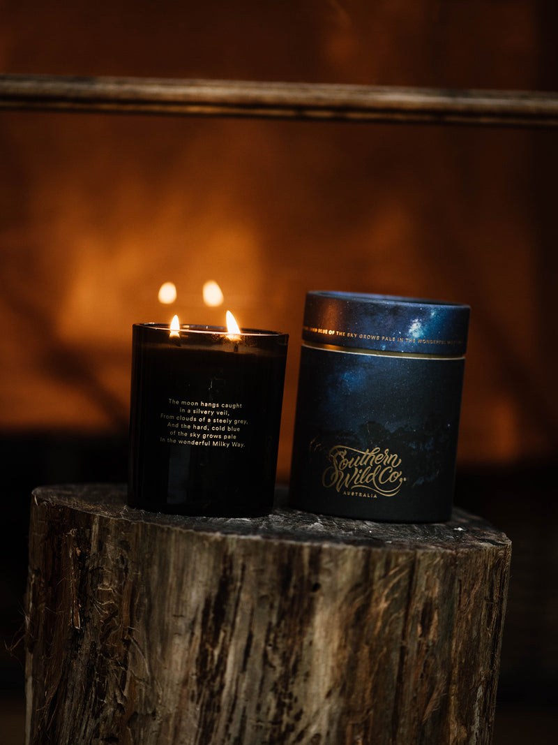 A lit Southern Sky candle and its celestial packaging rest atop a weathered tree stump, with the warm glow of a fire behind them—evoking winter rituals and the quiet magic of starlit nights.