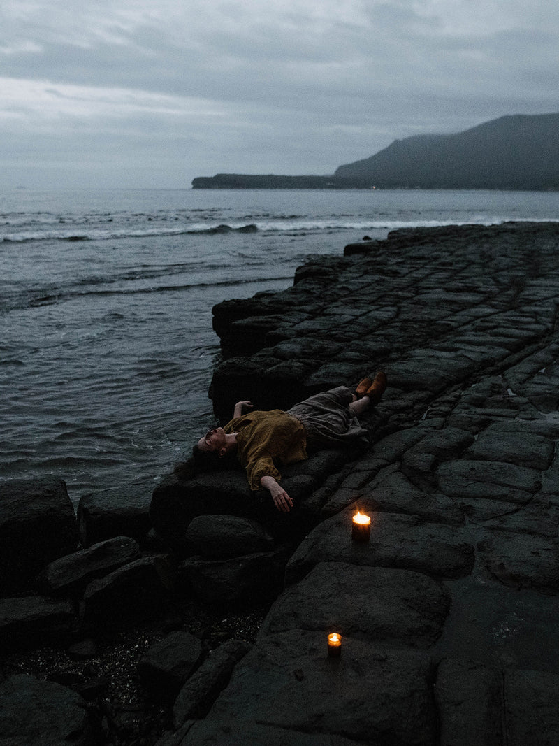 Elle-May Watson reclines on dark coastal rock as Southern Sky candles flicker beside her—evoking wild oceans, wide skies, and the serenity of stargazing in Australia.