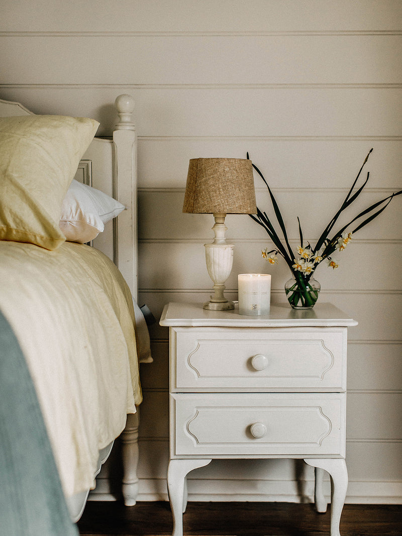 Sunshine candle glowing softly on a white bedside table, beside spring daffodils and a linen-shaded lamp — a gentle invitation to slow mornings and golden light.