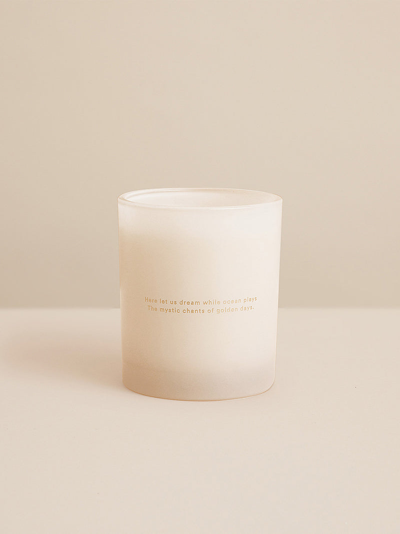 Single Sunshine candle photographed against a warm neutral backdrop, etched with poetic gold type — ‘Here let us dream while ocean plays.’