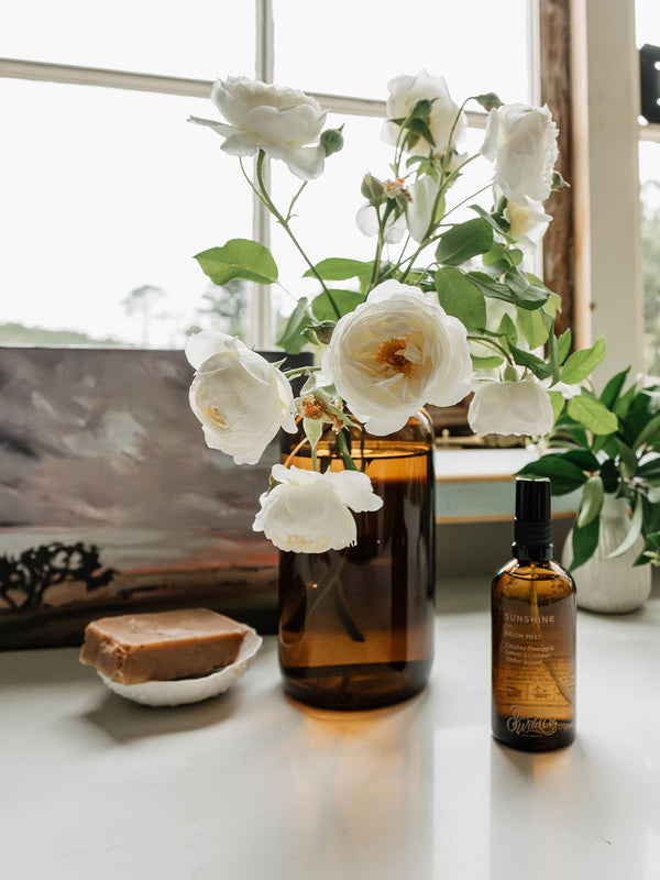 Sunshine Room Mist styled on a windowsill beside ivory garden roses and natural soap — a breath of summer light in an everyday ritual.