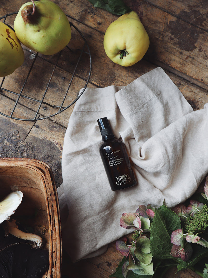 Amber bottle of Sunshine Room Mist resting on rustic linen with quinces, heirloom flowers and earthy textures — a nod to harvest, home and golden afternoons.