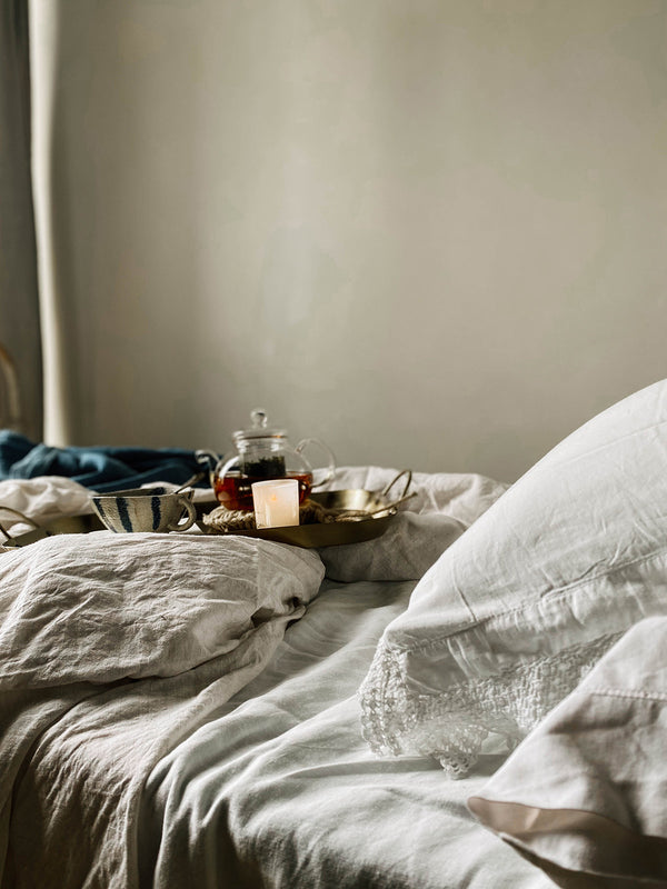 Sunshine votive candle glowing beside a teapot and ceramic bowl on a brass tray — a quiet morning ritual in bed, wrapped in linen and warmth.