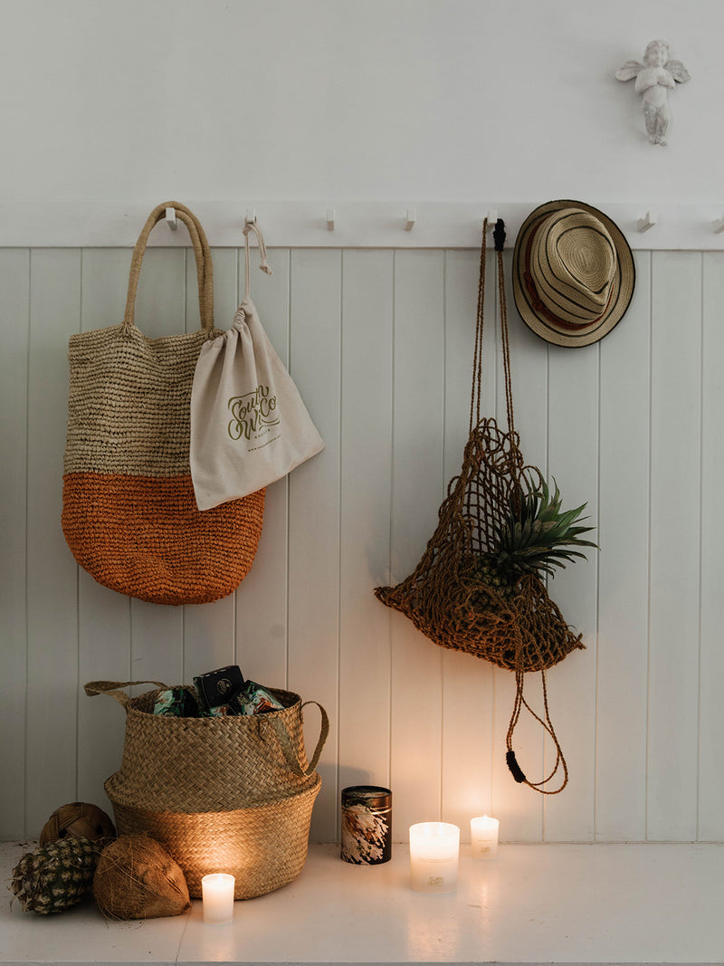 Entryway scene with glowing Sunshine votive and full-size candle among woven baskets, netted bags, and beach-foraged finds — a nod to sunlit days by the sea.