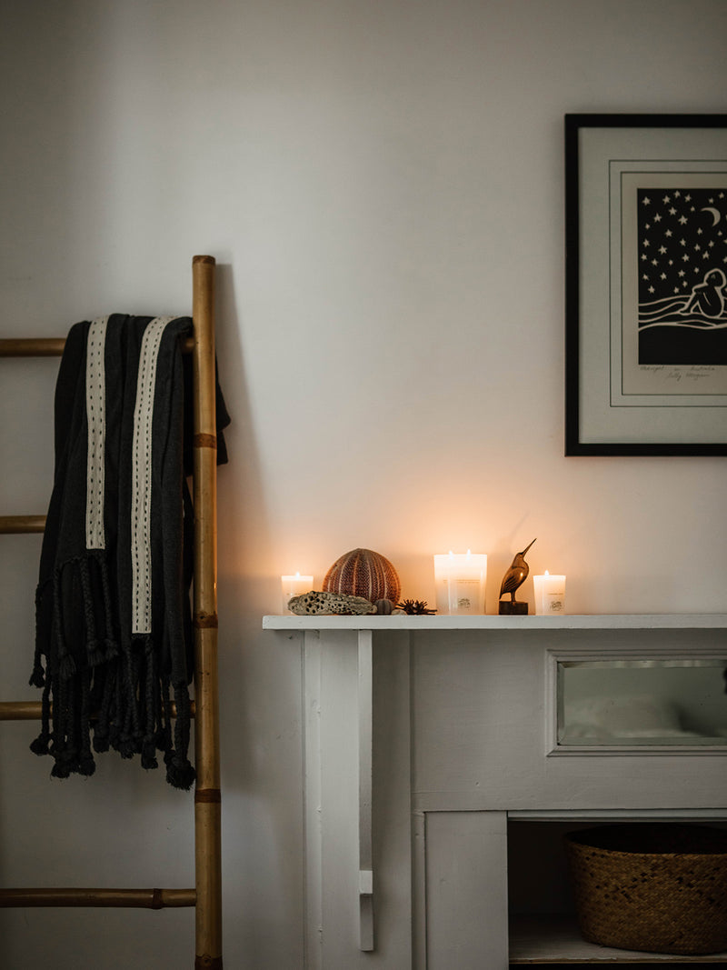 Lit Sunshine votive candle on a vintage white mantle, surrounded by coral, sea urchin shells and coastal treasures — soft light for a tranquil evening.