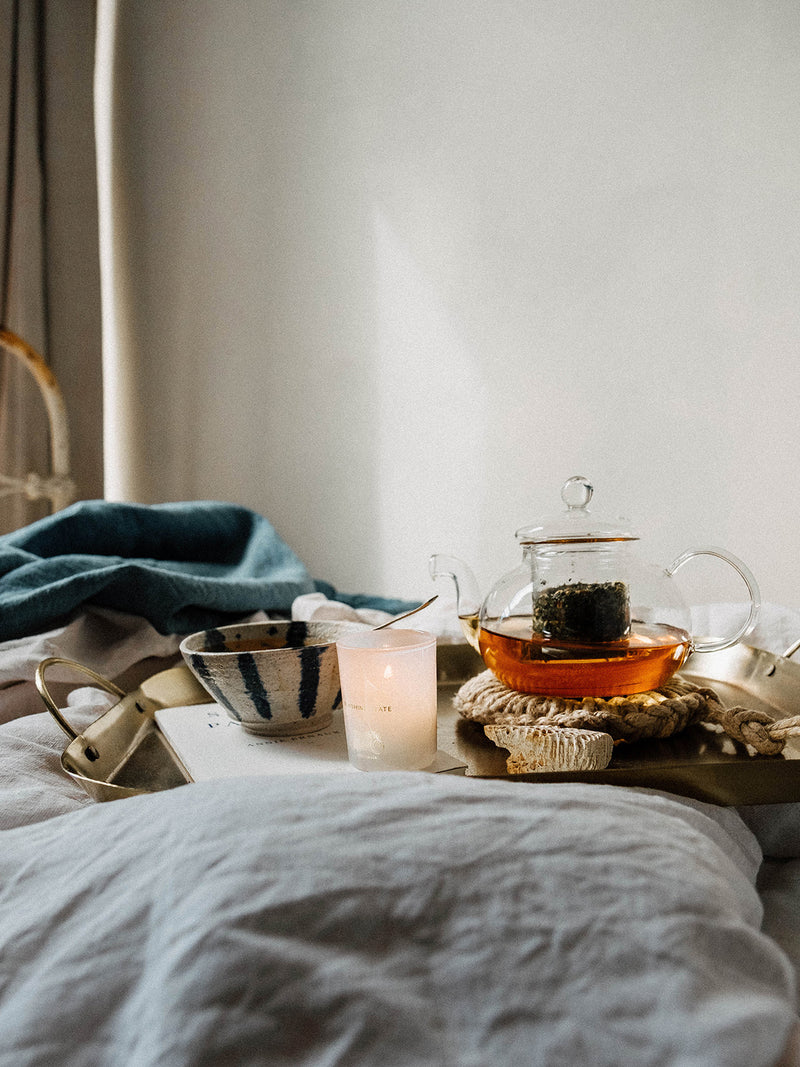 Softly lit bed scene with a brass tray holding tea, textiles, and a single Sunshine votive — a moment of stillness in golden morning light.