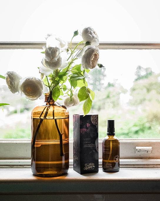 White roses sit in a vintage amber apothecary bottle next to a Southern Wild Co perfumed room spray on a bright and sunny window sill in a country style kitchen.