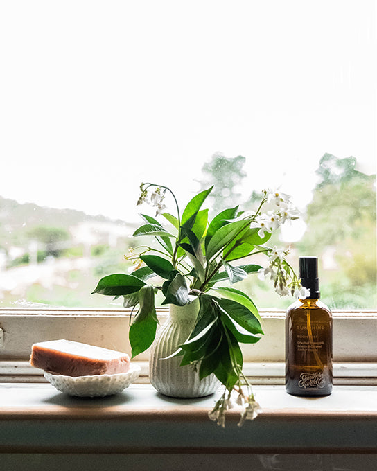 A pretty window sill in a country style kitchen. There are ceramics by Australian artists holding a natural soap and star jasmine flowers next to an amber apothecary bottle filled with perfumed room spray.